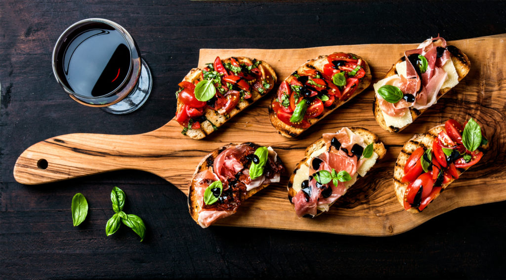 Brushetta set for wine. Variety of small saBruschetta sandwiches with prosciutto, tomatoes, parmesan cheese, fresh basil and balsamic creme served with glass of red wine on rustic wooden board over dark background, top view